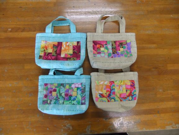 パッチワークの模様が中央にデザインされた、水色とベージュの小さな布製バッグが4つ並んでいる様子の写真