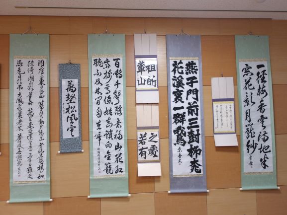 淡い色の布地に墨で書かれた縦長の書道作品がサイズや色が異なる状態で複数並べられて壁に展示されている様子の写真