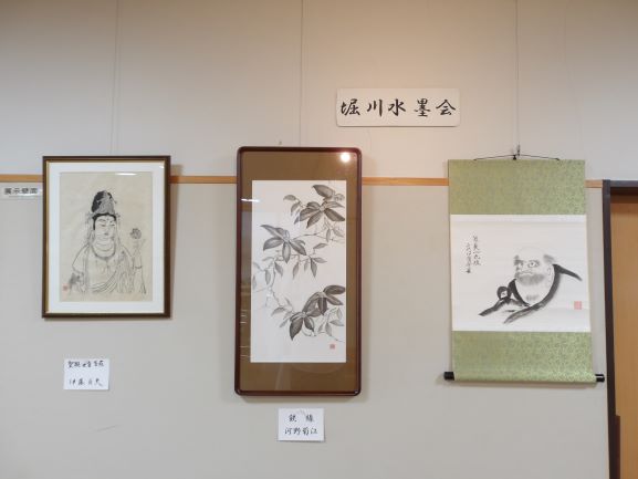 淡いベージュの壁に額縁に入った人物画と、葉のついた枝が描かれた水墨画が掛け軸とともに展示されている様子の写真