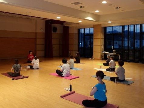 フローリングの広間の中で、数名の女性がヨガマットの上に座って講師の指導を受けながら、夜の窓の外を背にしてヨガを行っている様子を捉えた写真