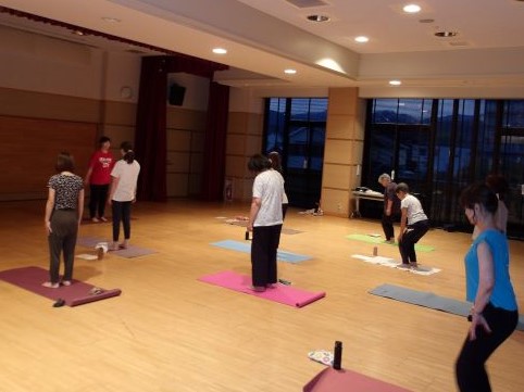 広い部屋で複数の男女が各自のヨガマットの上に立ち、夜の窓を背にして講師の指導を受けながらヨガのポーズをとっている様子を捉えた写真