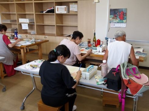 教室内の複数のテーブルで老若男女が各自の絵画制作に集中しており、奥の棚には様々な道具が並んでいる様子の写真