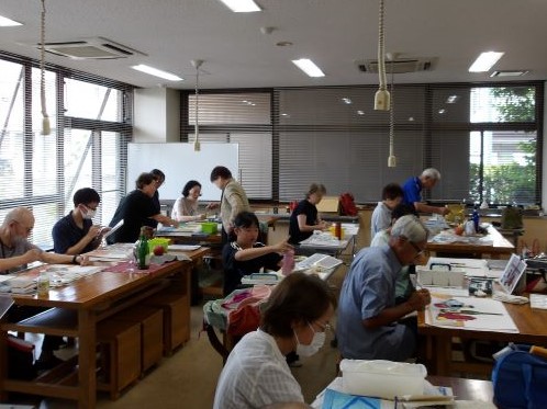 様々な年代の人々が大きな部屋でそれぞれテーブルについて、絵を描いたり作業をしたりと各自の活動に集中している様子の写真