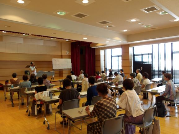 木目の床が広がる室内で、壇上の指導者がホワイトボードを前に、机と椅子に座る複数の生徒たちに対し説明している様子を捉えた写真