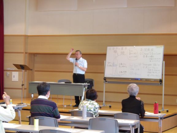 男性の指導者がホワイトボードを背に壇上に立ち、腕を上げて合唱や声楽を教えており、その教えを聴き入る複数の生徒が向かい合う写真