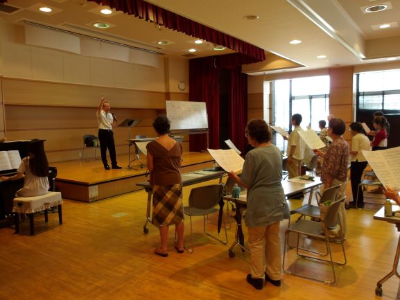 ホールの壇上で指揮をとる加藤さんの指導者と、手元の譜面を見ながら歌っている生徒たちを捉えており、ピアノを弾く女性も写る合唱の練習風景の写真