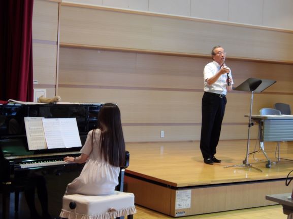 ホール内で、壇上の加藤さんがマイクを手に、ピアノを弾くロングヘアの女性の生徒に歌唱指導を行っている様子を捉えた写真