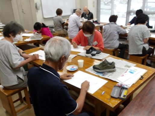 体育館のような広い部屋で、複数の高齢者が机に座り、靴を見本に描いている様子の写真