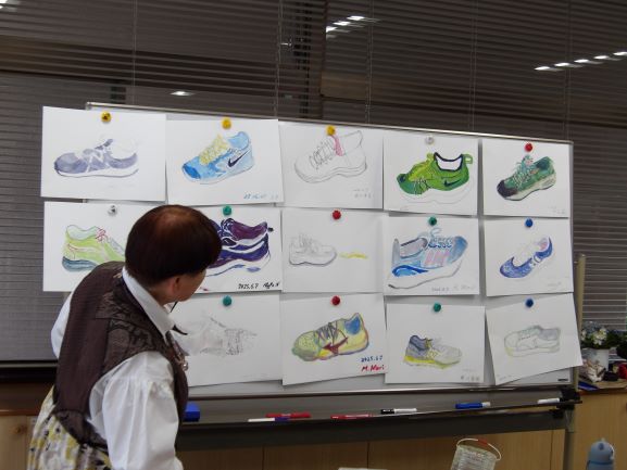 机に立てかけられたホワイトボードに、複数の靴のデッサンや水彩画がピンで留められており、それらを鑑賞している人物を写した写真