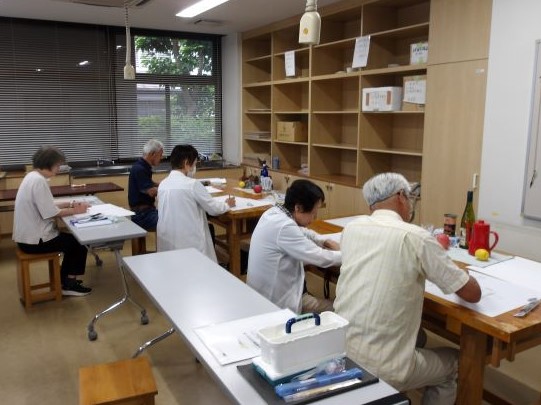 机に向かって複数の高齢者が静かにデッサンに取り組んでおり、室内には棚や窓がある絵画教室の様子の写真