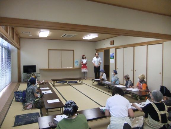 和室で、エプロンと三角巾を着用した男女が、座布団に座る複数の人々に何かの説明をしている様子を捉えた写真