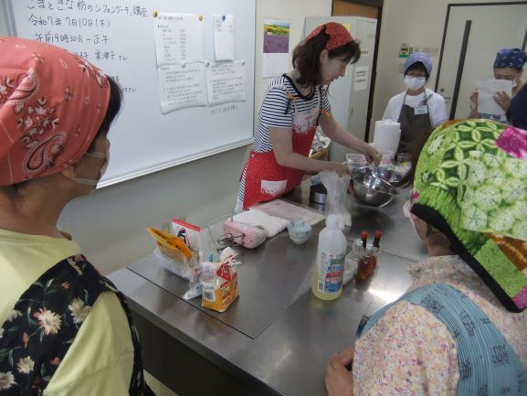 頭にバンダナを巻いた臼杵さんが、ステンレスの調理台で参加者たちにシフォンケーキ作りを教えながら、ボウルの中身を混ぜている様子を捉えた写真