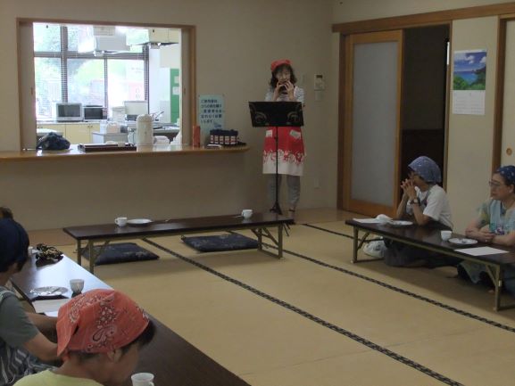 赤いエプロンとバンダナを身につけた女性がマイクを手に、座布団に座る参加者たちを前に何かを話している様子を捉えた写真