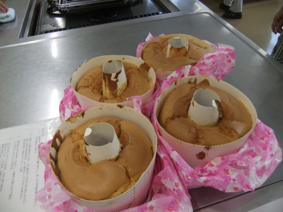 銀色の調理台に、紙でできた型に入った焼きあがりのシフォンケーキが4つ並べられており、底の紙がめくれあがってふわふわのスポンジが見えている写真