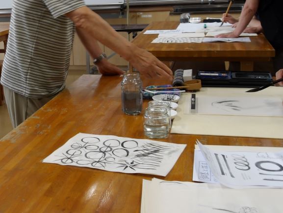 木製の机に墨と筆、水差しが置かれ、複数の人々が筆を手に、墨の濃淡を活かした様々な模様や線を練習している書道教室の風景を捉えた写真