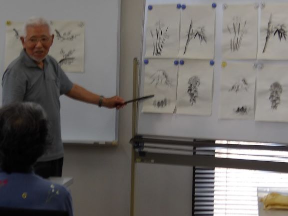 教室内で、笑顔の年配男性がホワイトボードに貼られた水墨画の作品を指し棒で指し示し、参加者に作品を説明している様子を捉えた写真