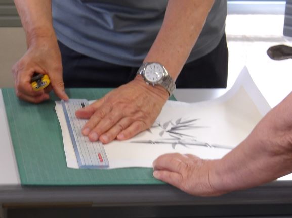 書道教室の机の上で、カッターと定規を使って墨で描かれた竹の絵を切り、その隣には別の人が手を添えて作業を手伝っている様子を捉えた写真