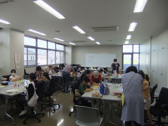 室内で複数の机を囲むようにたくさんの子供たちが座り、大人たちが指導をしながら各自の課題に取り組む、賑やかなワークショップの風景を捉えた写真