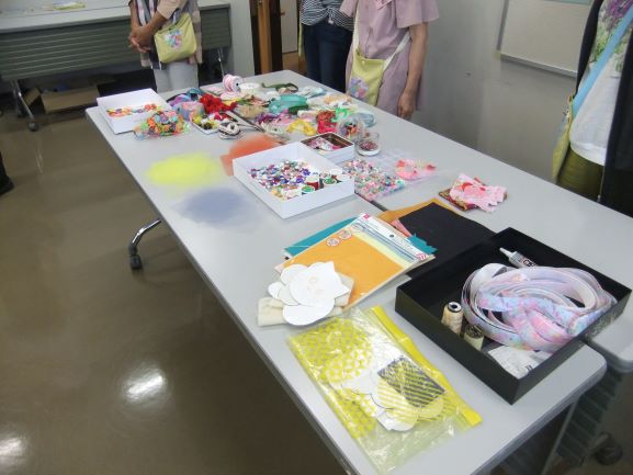 作業テーブルの上に、リボンや布、ビーズ、接着剤、型紙など、手芸の材料が所狭しと並べられており、後ろには立って見守る人々の足元が写る写真