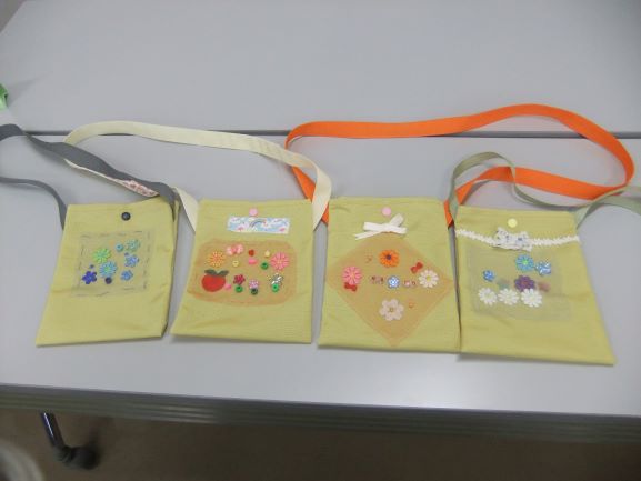 薄い黄色の布地にカラフルなビーズやフェルトの花、リボンなどで個性的に装飾された4つの手作りのミニバッグが並べられている写真