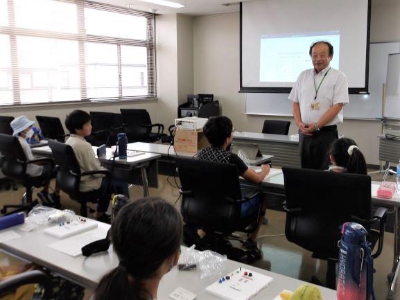 白いプロジェクタースクリーンの前で久保田さんが微笑みながら立ち、机に座った数人の子供たちに何かを教えている様子を捉えた写真