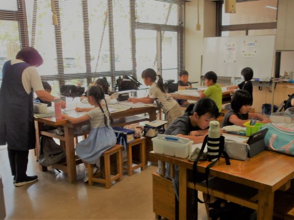 教室内で、複数の子供たちが机に向かってそれぞれ集中して作業しており、前田さんが立って生徒に指導している様子を捉えた写真