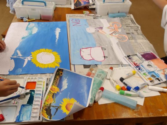 画板と水彩絵の具、筆が置かれた作業台で、子供たちがひまわりや自転車の絵を制作しており、横にはお手本となるひまわりの写真が置かれている写真