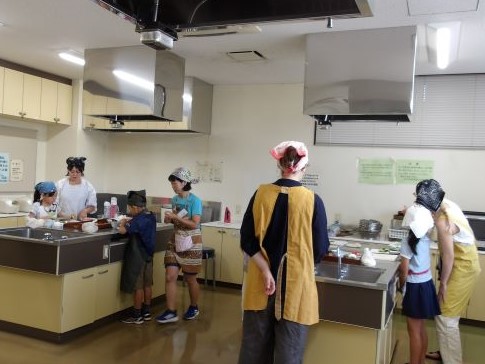 ステンレスの流し台が複数ある広い調理室で、エプロンや三角巾を身につけた子供たちが、大人たちの指導を受けながら調理に取り組んでいる様子を捉えた写真