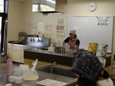 エプロンと三角巾を着用した先生らしき女性が作業台に向かい、別の参加者が手前のシンクのそばで作業をしている様子を捉えた写真