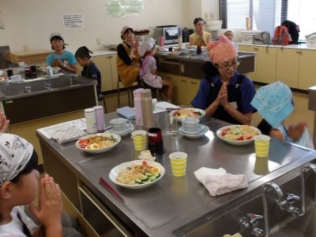 流し台や調理台が並ぶ部屋で、三角巾とエプロンをつけた子供たちや大人たちが、自分たちで作った料理を前に手を合わせて「いただきます」と挨拶している写真