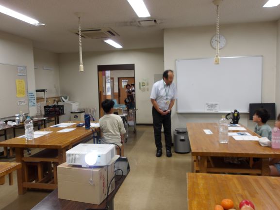 子供たちに対し、久保田さんがホワイトボードのそばに立って何かを話している様子を捉えた写真