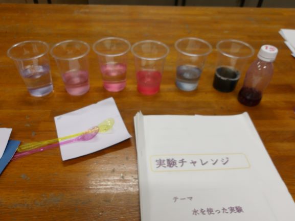 木製のテーブルの上に「実験チャレンジ」と書かれた冊子と、色の異なる液体が入った複数の透明なプラスチックコップが並んでいる様子を捉えた写真