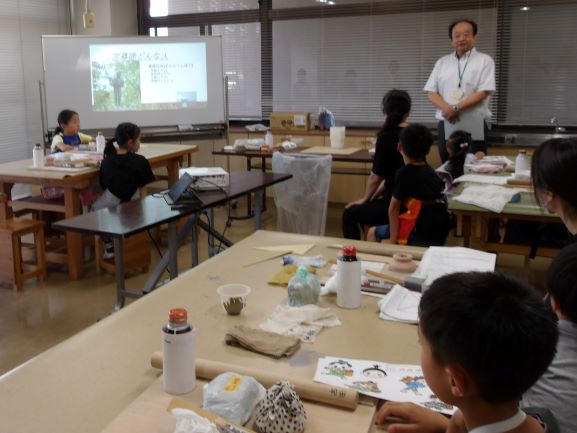 室内の長机を複数並べた空間で、男性がプロジェクターのスクリーンを前に立ち、数人の子供たちに囲まれながらペン立て作成を教えている場面を捉えた写真