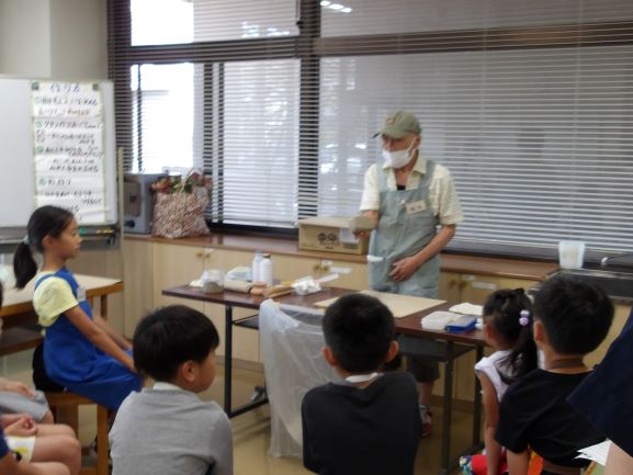 エプロンと帽子を着用したマスク姿の年配の男性が、机を囲む複数の子供たちに作品を見せながら指導している風景を捉えた写真