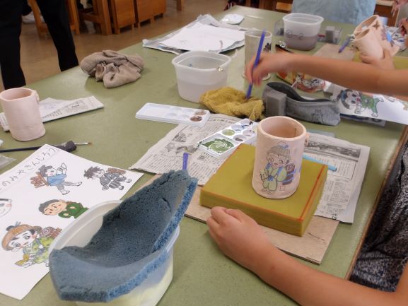 木目調の作業台で、子供たちが陶芸作品に彩色しており、紙に描かれたキャラクターのイラストや絵の具、水入れが周囲に散乱している様子を捉えた写真