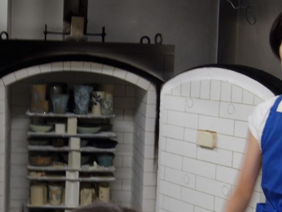 素焼きの器やコップを複数段の棚に並べて窯の中に入れており、その隣で作業着を着た人が立っている様子を捉えた写真