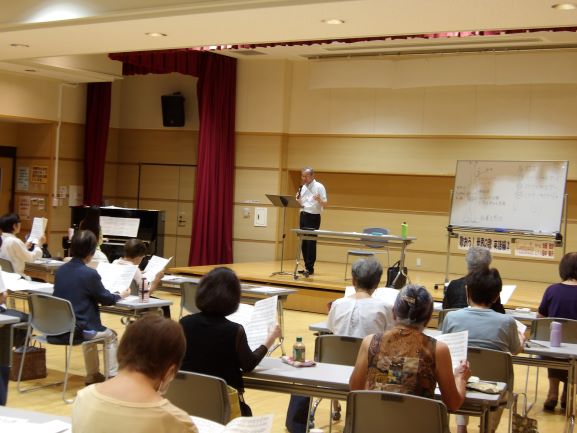 広々とした室内で、男性の指導者が壇上に立ち、譜面台とホワイトボードを前に椅子に座る十数名の生徒たちに合唱の指導をしている写真