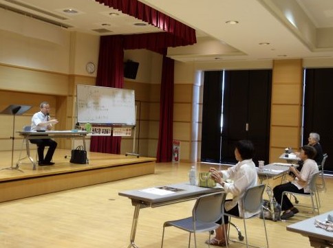 広々としたホールで、指導者が壇上の机に座り、前方で机に向かい座る数名の生徒に合唱の指導を行っている写真