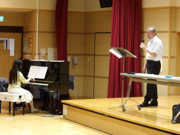 ステージ下で黒いアップライトピアノを弾いている女性と、ステージ中央でマイクを片手に歌を歌っている様子の男性の写真