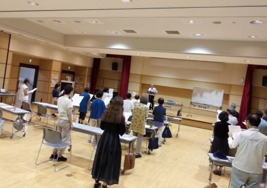 ホールの中で、男性の指導者が壇上から向き合い、譜面台と机を前に並び、合唱の練習に励む複数の人々を捉えた写真