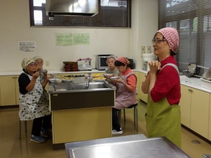 調理実習室でエプロンとバンダナを着用した数人の女性たちが、講師の話を聞いており、中央のステンレス製シンク周りでは数人が調理や配膳を行っている様子の写真