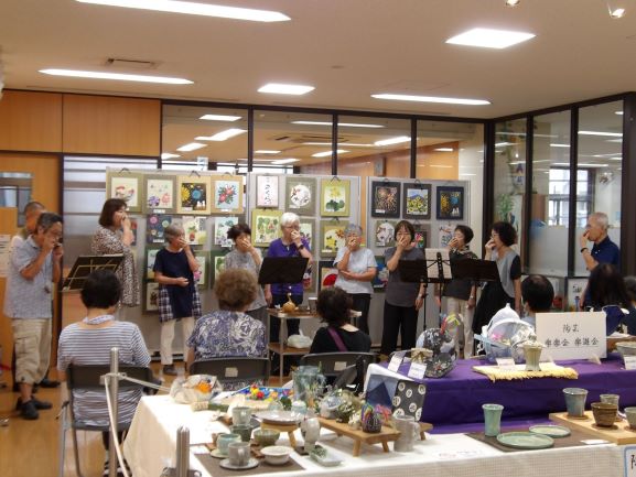 屋内の一室で、数人の演奏者が口元に手を当てて楽器を演奏し、その後ろの壁には色鮮やかな絵画作品が多数展示され、手前のテーブルには陶芸作品が並べられている写真