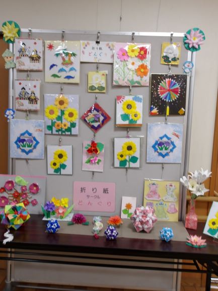 白い展示パネルとテーブルの上に、花をモチーフにした作品が多数飾られており、パネルにはひまわりや花などの色鮮やかな折り紙が飾られている写真