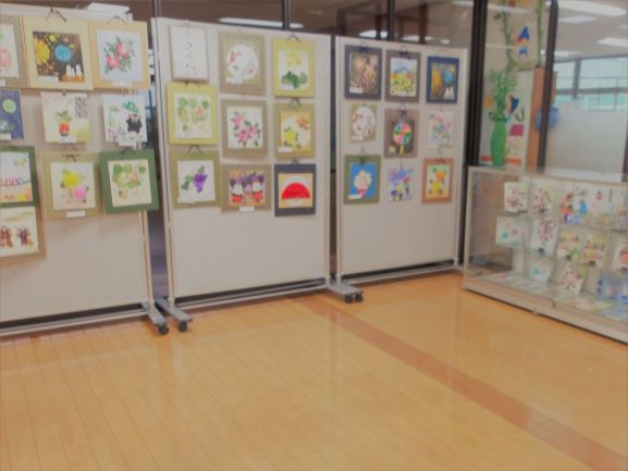 明るいフローリングの室内の展示スペースに、白いパネルが立てられ、そこには花や動物、風景など様々なモチーフをカラフルに描いた絵画作品が多数飾られている展示会場の様子の写真