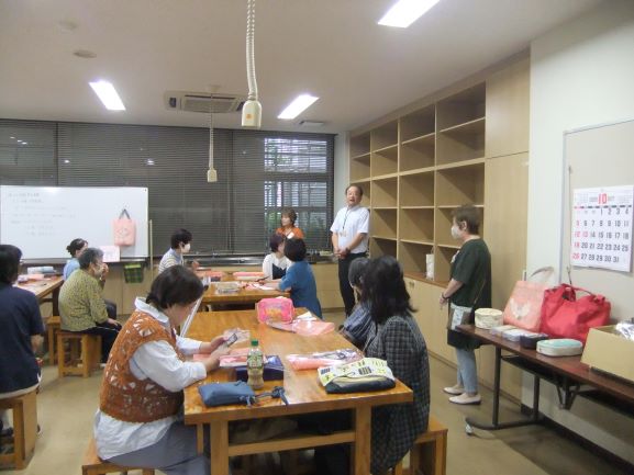 室内で、多くの参加者が木の机を囲んで座り、中央に立つ白いシャツの男性の話を笑顔で聞いている様子の写真