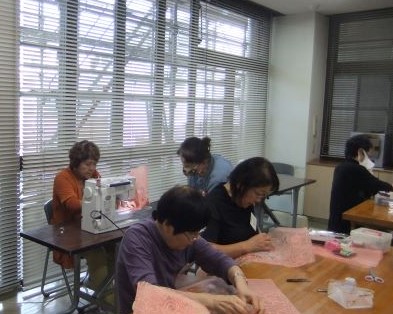 大きな窓から光が差し込む明るい室内で、数人の女性たちがテーブルを囲んで座り、一人がミシンを使い、他の参加者たちは薄いピンク色のレース生地を手作業で縫い合わせるなど、手芸活動に取り組んでいる様子の写真