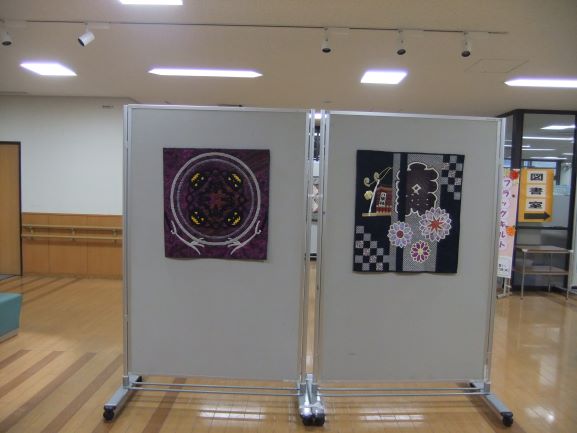 明るいフローリングの室内に設置された白い展示パネルに掛けられた二つのパッチワークキルト作品であり、左は紫を基調としたマンダラのような幾何学模様で、右は黒地に花や家などのモチーフが配置された和風の作品の写真