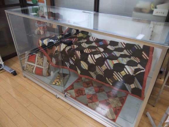 ガラスケースの中に展示された手芸作品群であり、黒い生地に様々な色の布を斜めに縫い付けた大きなパッチワークキルトと、赤とグレーの格子柄の小さなパッチワークのバックが飾られている写真