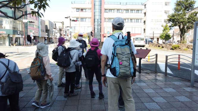 晴れた日の市街地にある交差点の舗装された広場で、リュックサックを装備した参加者のグループが集合して説明を聞いている様子の写真