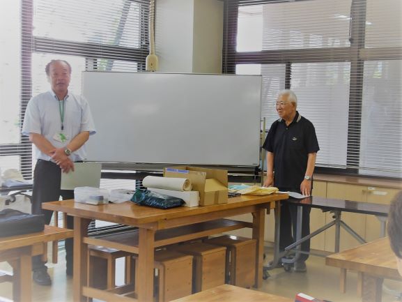 部屋の中で、ホワイトボードを背に机を挟んで二人の男性が立ち、参加者達を前に説明している様子を捉えた写真